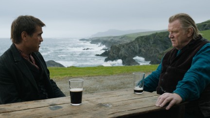 With mountains and sea in the background, an older man sits at a table with a nearly empty pint of Guinness in front of him, looking at a younger man who has an upset expression on his face. He has a half pint of Guinness in front of him. 